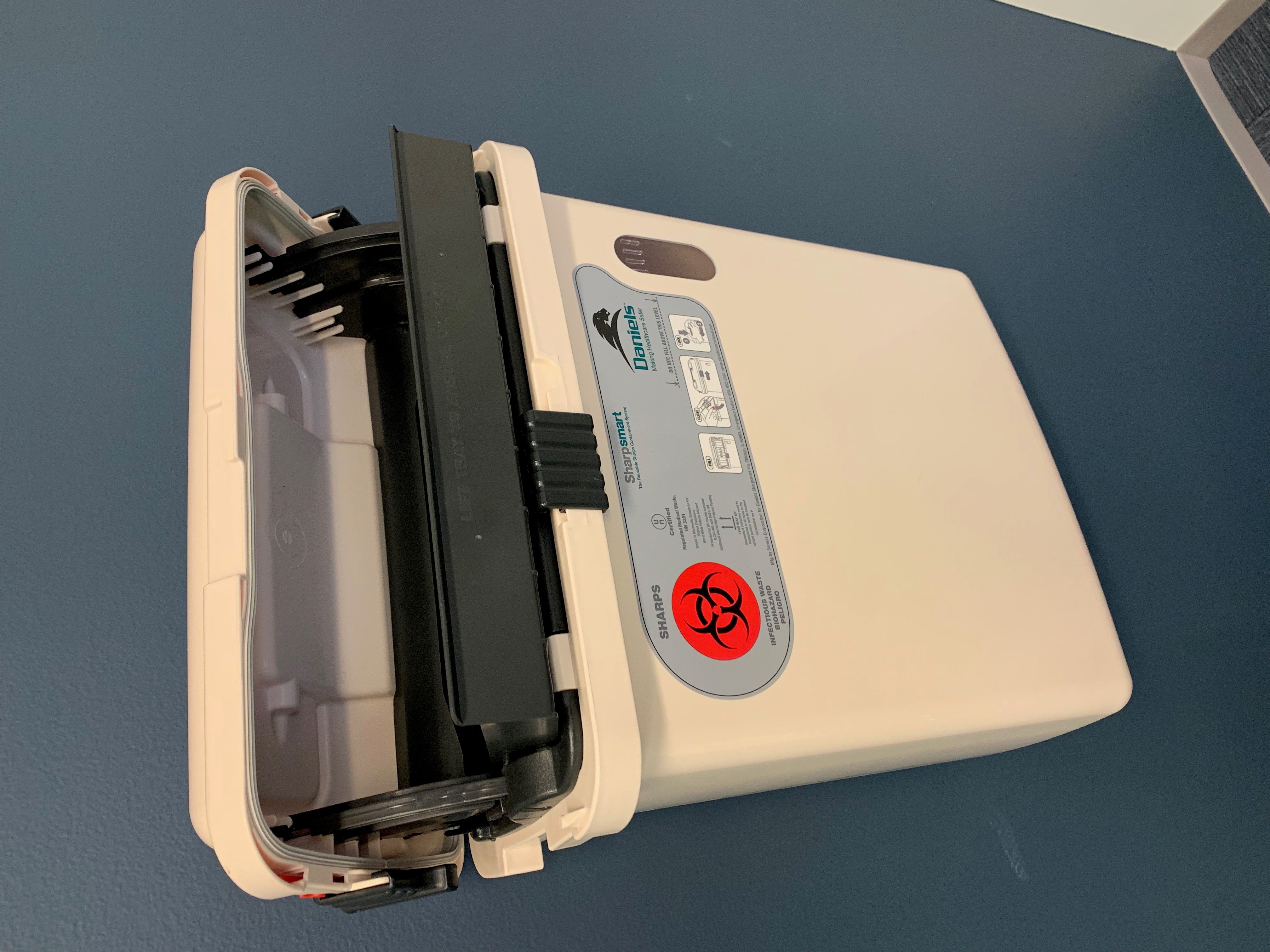 A puncture-resistant, leak-proof sharps container. Relying on mechanics and gravity, used needles must be deposited immediately into this bin without the risk of two-handed recapping.
